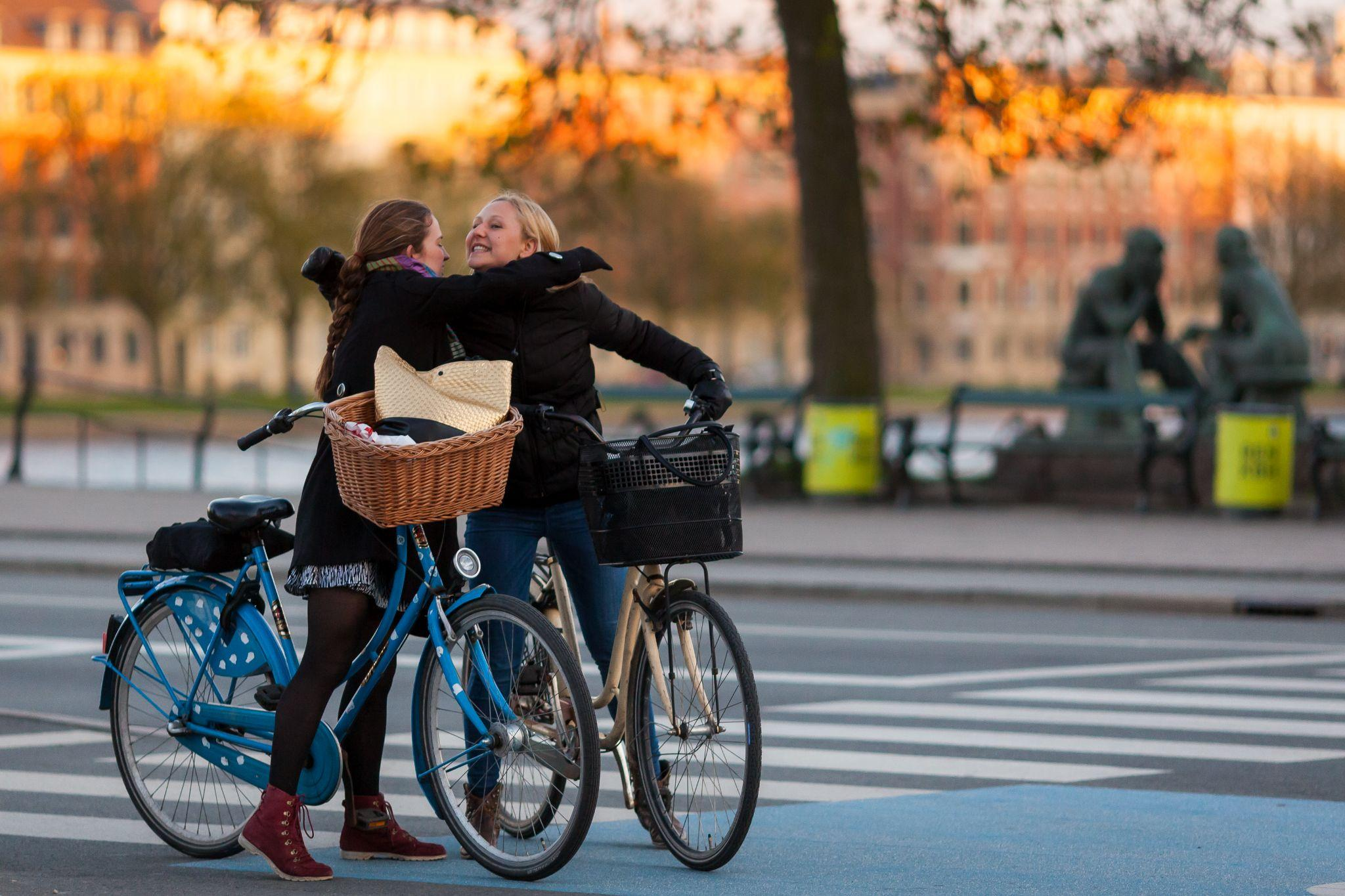 コペンハーゲンの街かどで二人の女性が自転車にまたがったまま楽しそうにお互いの左肩に右腕を載せています(写真)