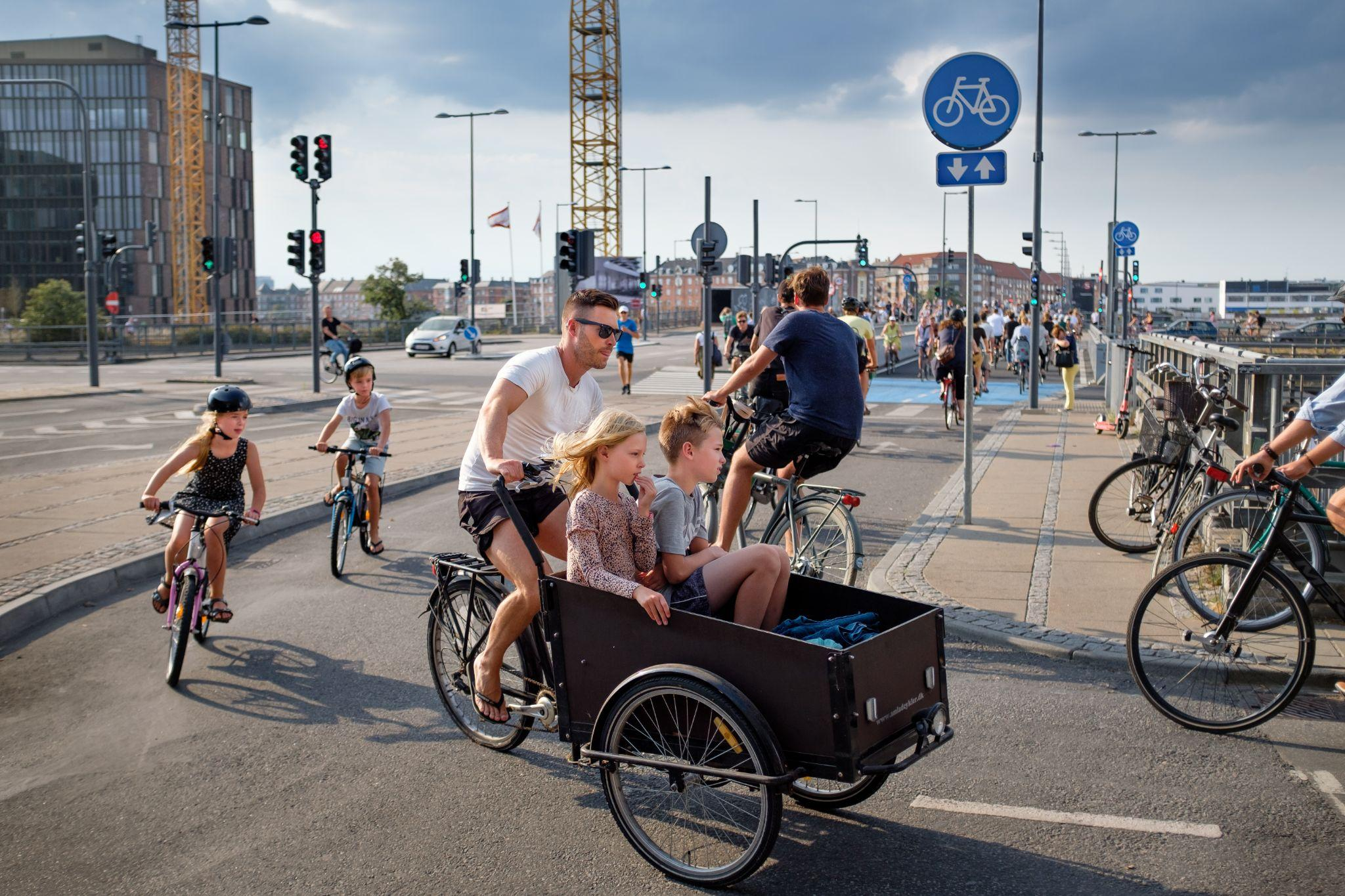 コペンハーゲンの自転車専用レーンで、前にリアカーのような大きな荷台のある自転車に子ども2人を乗せて父親と思われる人が漕いでいます(写真)